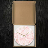 Horloge murale ronde marbre rose et or emballée dans sa boîte de rangement en carton sur fond de bois sombre.