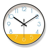Horloge murale nordique au design scandinave, avec cadran blanc et partie inférieure jaune moutarde. Les aiguilles noires contrastent avec les chiffres bleus. Son contour peut être noir ou doré. Cette horloge murale moderne et élégante apporte une touche chaleureuse et originale à un salon, une chambre ou un bureau