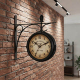 Une horloge murale double face noire, de style industriel inspiré des gares, est fixée à un mur gris au-dessus d'une console. L'horloge a un cadran de couleur crème avec les chiffres romains I, II, III, etc. et la mention "New York". En arrière-plan, on voit un salon avec un canapé gris, une bibliothèque en bois et une plante verte.
