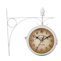 horloge murale double face identique à celle de l'image 0.png, mais avec un support ornemental en métal peint en blanc. Le cadran crème vieilli, les chiffres arabes noirs et le texte "GRAND CENTRAL TERMINAL - NEW YORK" sont clairement visibles. L'horloge est fixée sur un fond blanc uni.