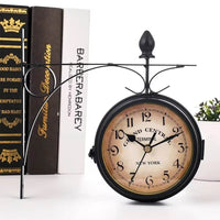 L'horloge murale double face de style vintage, avec son support en métal noir, est posée sur une surface blanche. Derrière elle, sur la gauche, se trouvent trois livres empilés verticalement. Le livre du bas est noir avec des détails dorés, le livre du milieu est noir avec une tranche dorée, et le livre du haut est blanc avec le titre "BAREARABAREY" en noir. Sur la droite, une petite plante en pot vert est posée sur la surface blanche. Le cadran de l'horloge est crème vieilli avec des chiffres arabes noirs 