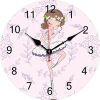 Une horloge murale ronde au fond rose poudré décoré de motifs floraux discrets. Au centre, une illustration montre une jeune ballerine en tutu blanc et violet avec une couronne de fleurs. Les chiffres noirs lisibles et les aiguilles noires et rouge complètent le cadran