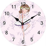 Une horloge murale ronde au fond rose poudré décoré de motifs floraux discrets. Au centre, une illustration montre une jeune ballerine en tutu blanc et violet avec une couronne de fleurs. Les chiffres noirs lisibles et les aiguilles noires et rouge complètent le cadran