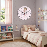 L'horloge murale ronde danseuse est fixée au mur mauve clair d'une chambre de petite fille. La pièce est décorée dans des tons doux avec un lit en bois, une couette à rayures roses, des peluches et une bibliothèque garnie de livres pour enfants