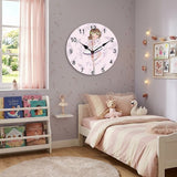 L'horloge murale ronde danseuse est fixée au mur mauve clair d'une chambre de petite fille. La pièce est décorée dans des tons doux avec un lit en bois, une couette à rayures roses, des peluches et une bibliothèque garnie de livres pour enfants