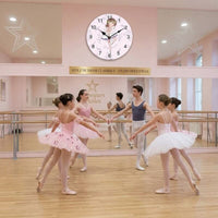 L'horloge murale thème ballerine est accrochée au mur d'un studio de danse classique au-dessus d'un grand miroir. Au premier plan, de jeunes danseurs en tutu rose et tenues de ballets répètent au centre de la salle au parquet clair