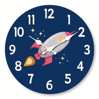 Horloge murale fusée spatiale avec une fusée grise et rouge bordeaux au centre du cadran. Les flammes donnent une impression de mouvement et de puissance. Des étoiles entourent la fusée sur un fond bleu foncé. Les chiffres et les aiguilles blanches ressortent clairement.