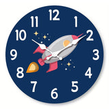 Horloge murale fusée spatiale avec une fusée grise et rouge bordeaux au centre du cadran. Les flammes donnent une impression de mouvement et de puissance. Des étoiles entourent la fusée sur un fond bleu foncé. Les chiffres et les aiguilles blanches ressortent clairement.