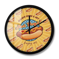 Horloge murale hot dog originale avec un sandwich au centre du cadran. Le hot dog est accompagné de ketchup et de moutarde. Le mot « Hot Dog » apparaît au-dessus. Chaque heure est représentée par un petit hot dog. Les aiguilles noires complètent ce design gourmand inspiré de la cuisine américaine.