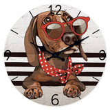 Horloge murale teckel représentant un adorable chien couché avec la tête posée sur ses deux pattes avant. Le teckel porte de grandes lunettes rouges originales ainsi qu'un foulard rouge à pois blancs qui lui donne un style unique et plein de charme. Son regard doux et attentif semble montrer toute sa tendresse et sa complicité avec son propriétaire. Le cadran blanc gris clair apporte une touche élégante tandis que les quatre bandes noires créent un design moderne qui met en valeur l'illustration du chien. 