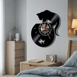l'horloge vinyle chat et bocal fixée sur un mur texturé gris au-dessus d'une table de chevet en bois. La scène inclut une lampe allumée, un réveil et un coin de lit, créant une atmosphère de chambre douce.