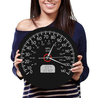 fille qui tient en mains l'Horloge murale ronde au design de compteur de vitesse automobile, dotée d'un fond noir mat, de chiffres et graduations blancs allant de 0 à 140, des mentions « MPH » et « km/h », d'un faux écran numérique à cristaux liquides affichant des chiffres gris, ainsi que d'aiguilles blanches et d'un axe central cerclé de rouge.