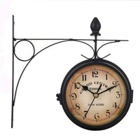 horloge murale double face de style vintage, fixée à un support ornemental en métal noir sur un fond blanc uni. Le cadran de l'horloge est de couleur crème vieilli, avec des chiffres arabes noirs de 1 à 12 et des aiguilles noires. Le texte 