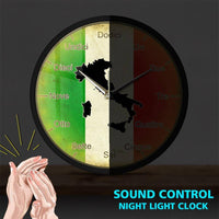 Horloge murale ronde italienne dans le noir, montrant le rétroéclairage LED activé. En bas à gauche, une illustration de mains qui applaudissent avec des rayons d'impact jaunes. En bas à droite, le texte en anglais "SOUND CONTROL NIGHT LIGHT CLOCK" en lettres bleues.