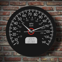 Horloge murale ronde au design de compteur de vitesse automobile, dotée d'un fond noir mat, de chiffres et graduations blancs allant de 0 à 140, des mentions « MPH » et « km/h », d'un faux écran numérique à cristaux liquides affichant des chiffres gris, ainsi que d'aiguilles blanches et d'un axe central cerclé de rouge sur mur brique