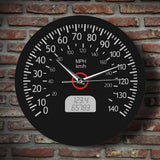 Horloge murale ronde au design de compteur de vitesse automobile, dotée d'un fond noir mat, de chiffres et graduations blancs allant de 0 à 140, des mentions « MPH » et « km/h », d'un faux écran numérique à cristaux liquides affichant des chiffres gris, ainsi que d'aiguilles blanches et d'un axe central cerclé de rouge sur mur brique