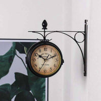 L'horloge murale double face de style vintage, avec son support en métal noir, est fixée à un mur blanc à l'intérieur d'une pièce. En arrière-plan, sur la gauche, un grand cadre noir contient une affiche représentant des feuilles de monstera vertes. Le cadran de l'horloge est beige avec des chiffres romains noirs et le texte "GRAND CENTRAL TERMINAL - NEW YORK".